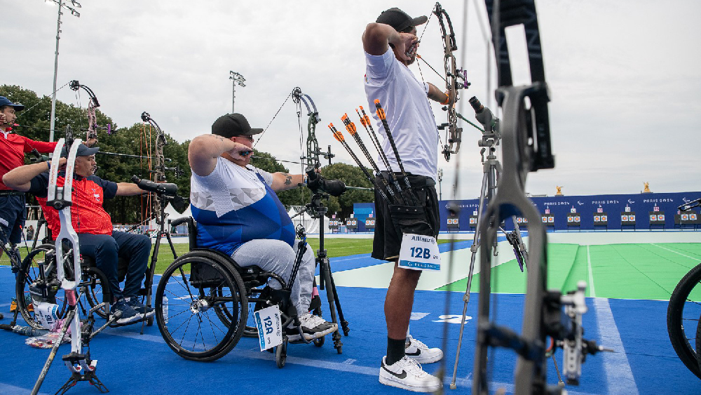 Jere Forsberg paralympialaisissa ampumassa jousiaseella kilpakumppaneidensa vierellä. Kuva otettu takaviistosta.